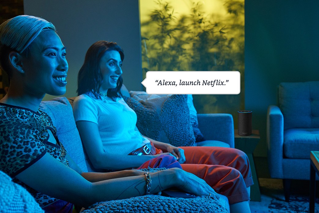 Two women watching TV, with a speech bubble over one saying, 'Alexa, launch Netflix'